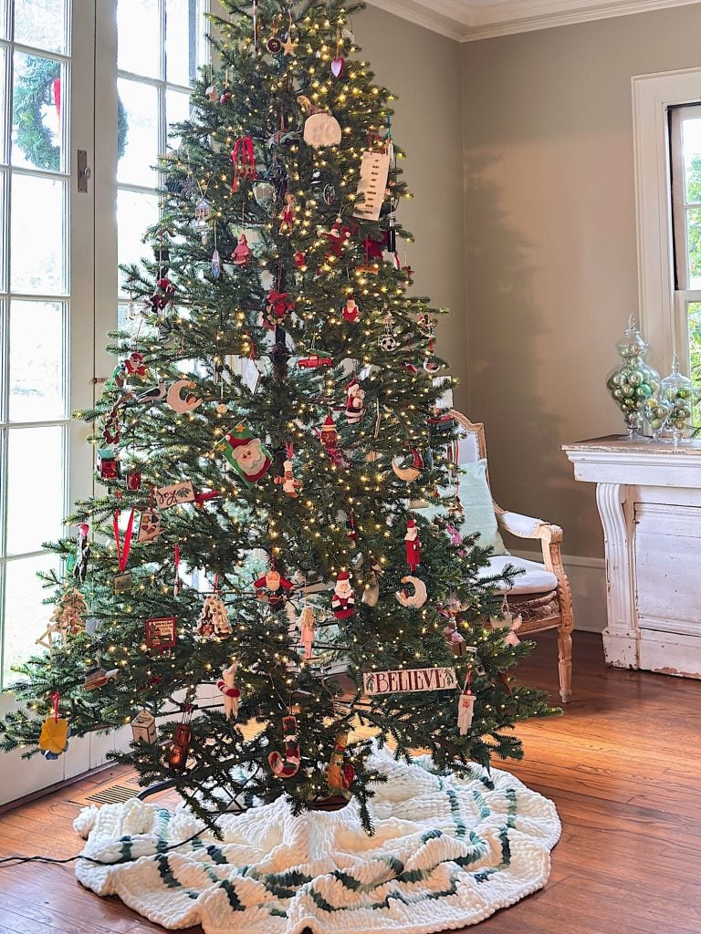A decorated Christmas tree with lights and ornaments stands next to a chair and a window in a bright living room with wooden floors.