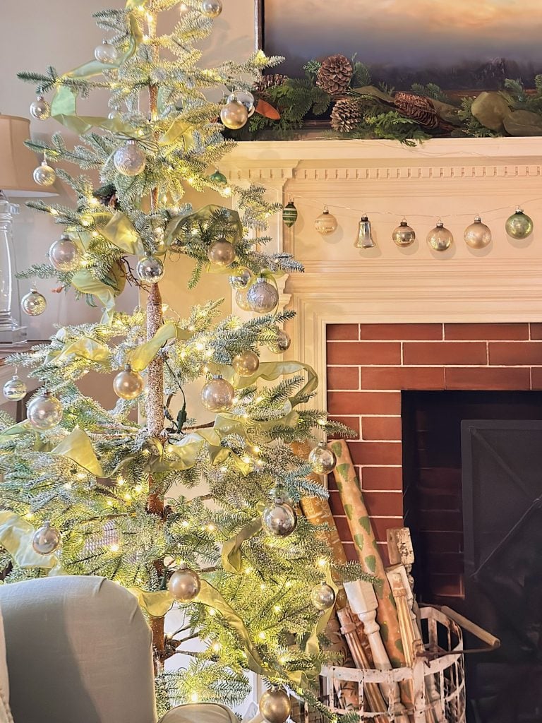 A decorated Christmas tree with silver ornaments and lights stands next to a fireplace with wrapped gifts in a basket and garlands on the mantel.