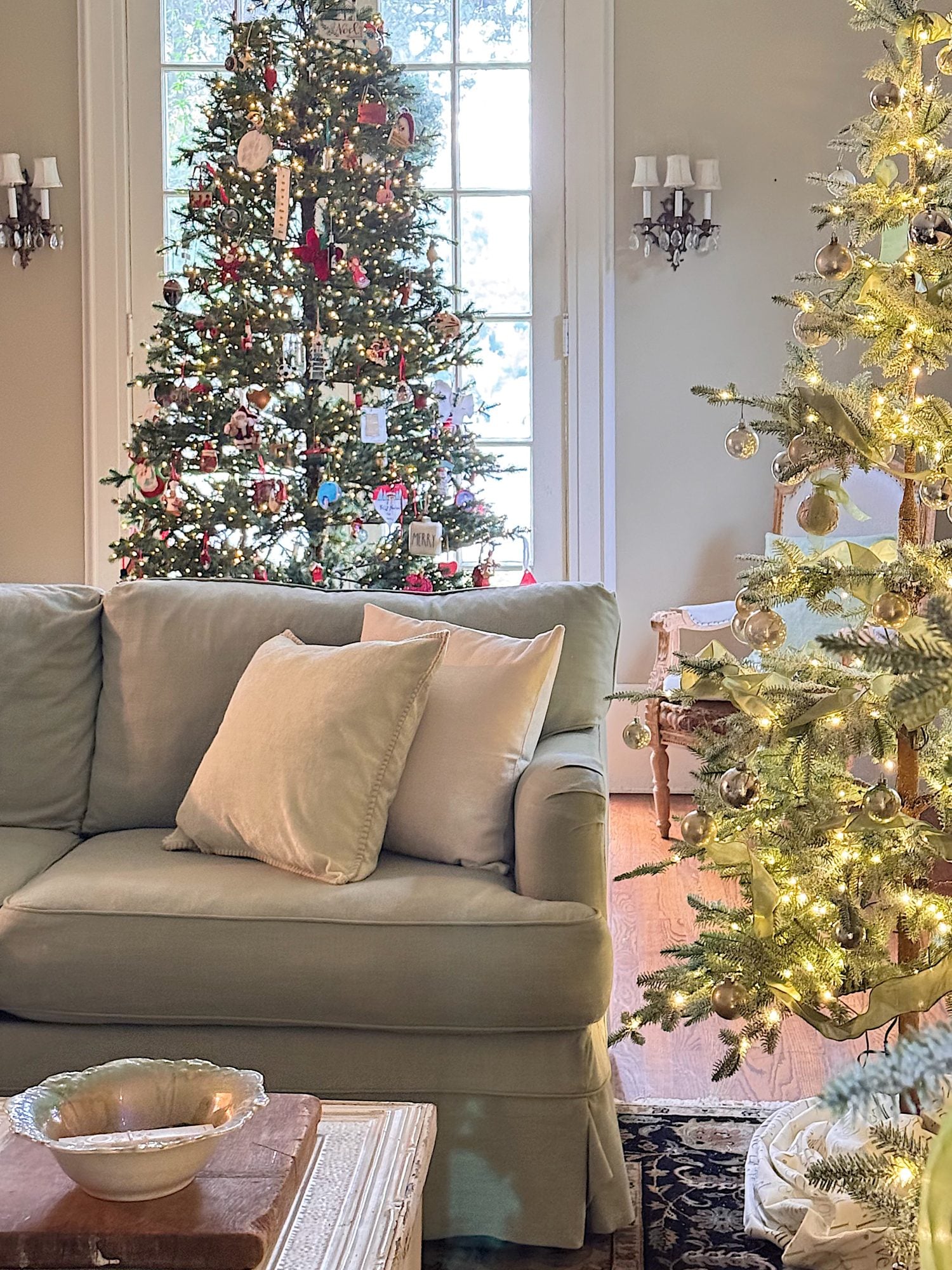 A living room with a light gray sofa and two decorative pillows, featuring two decorated Christmas trees near a window and natural light streaming in.