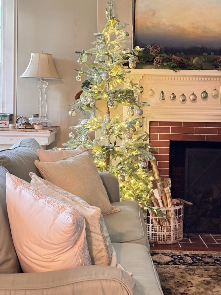 A small, decorated Christmas tree with white lights stands by a fireplace in a cozy living room with a sofa and pillows.