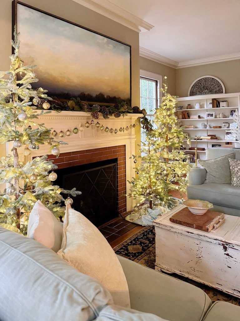 Living room with lighted Christmas trees, neutral sofas, a white distressed coffee table, wall art above the mantel, and built-in bookshelves in the background.