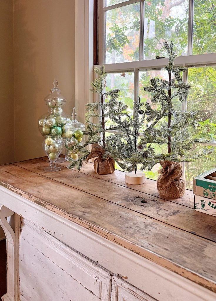 Two small artificial pine trees and jars filled with green ornaments are displayed on a rustic wooden table in front of a window.