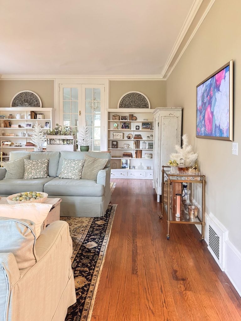 A living room with light-colored sofas, wooden floor, built-in shelves with books and decor, a bar cart, and a wall-mounted abstract painting.