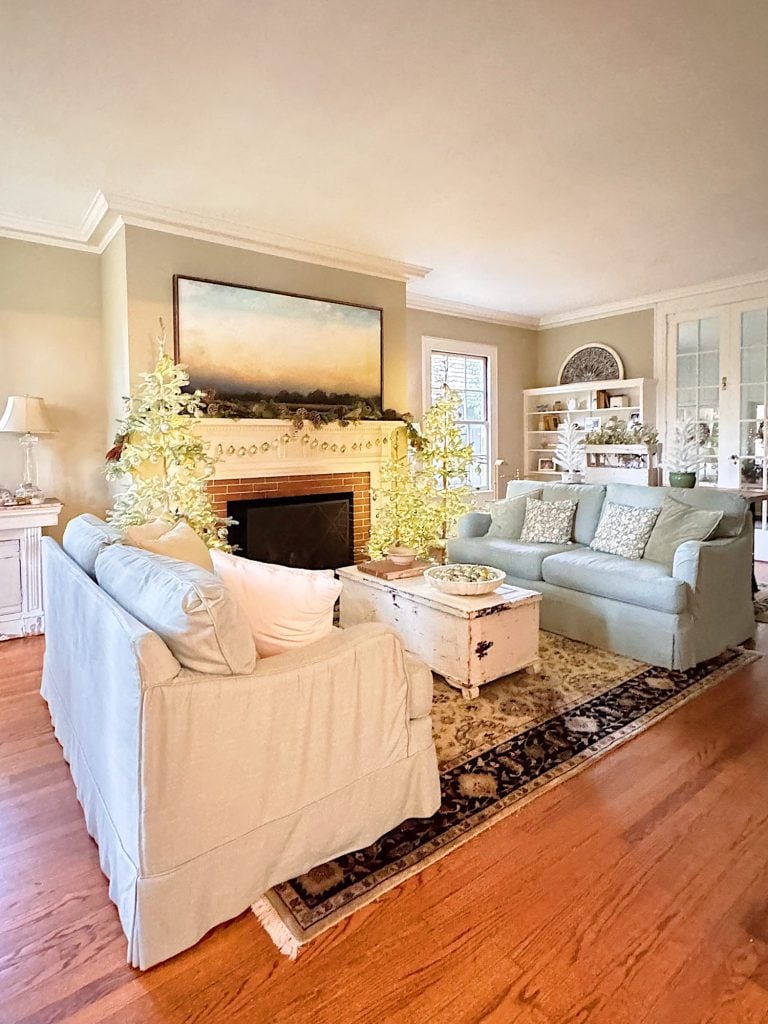 A bright living room with two light blue sofas, a white coffee table, a fireplace with decorative trees, and built-in shelves on hardwood flooring.