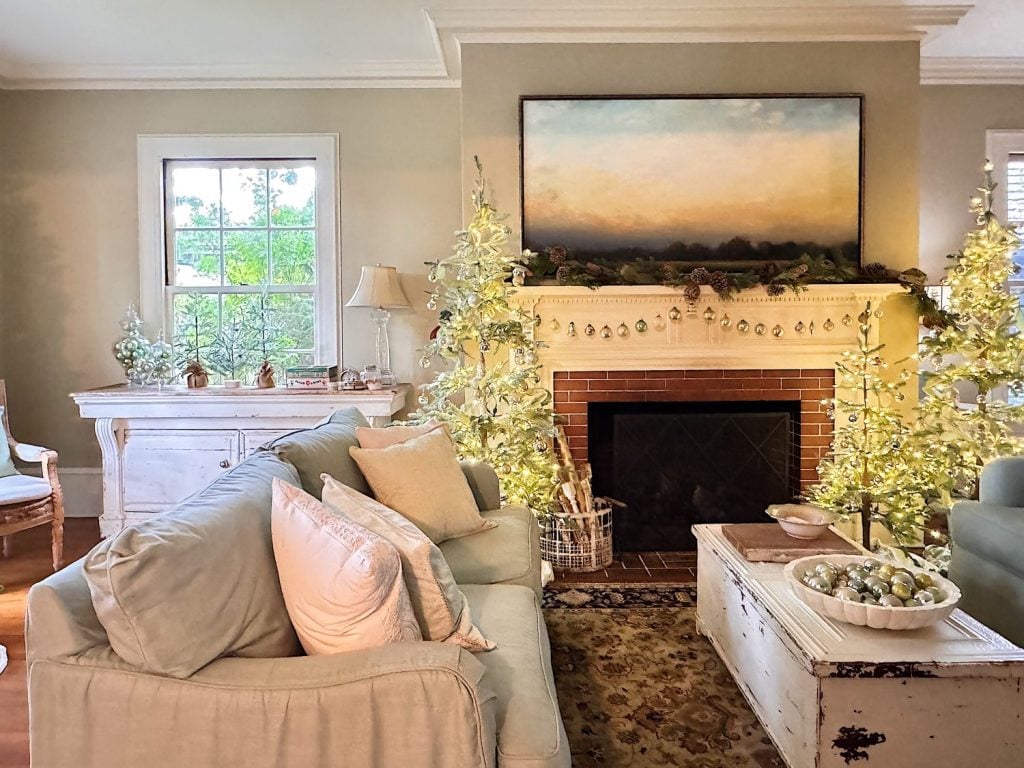 A cozy living room with light-colored furniture, a fireplace decorated with lit artificial trees, a landscape painting above the mantel, and a window letting in natural light.