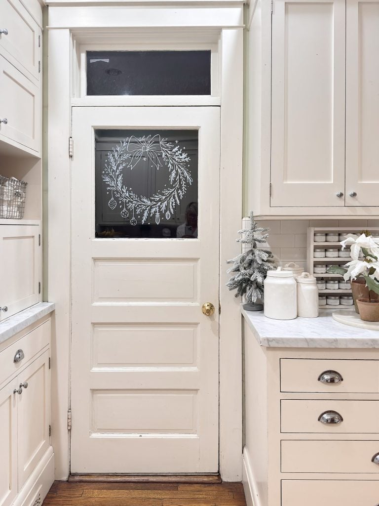White kitchen with a closed door featuring a drawn wreath on its glass panel; nearby are kitchen cabinets, countertop, small decorative tree, and paper towels.