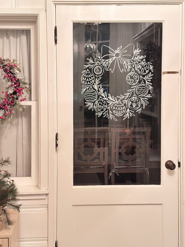 Glass door with a white line drawing of a holiday wreath featuring ornaments and a bow; holiday decorations are visible on a nearby window and part of a Christmas tree is in view.