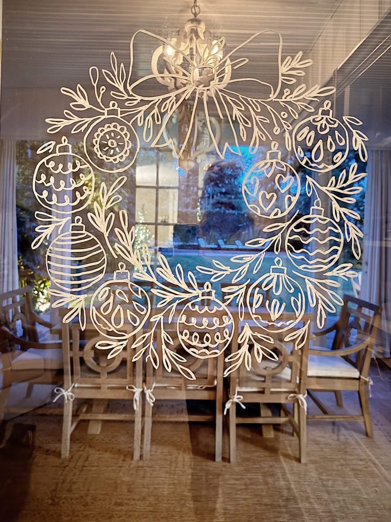 White line art of a holiday wreath with ornaments is drawn on a glass door, revealing a dining area with wooden chairs and a window view of trees outside.