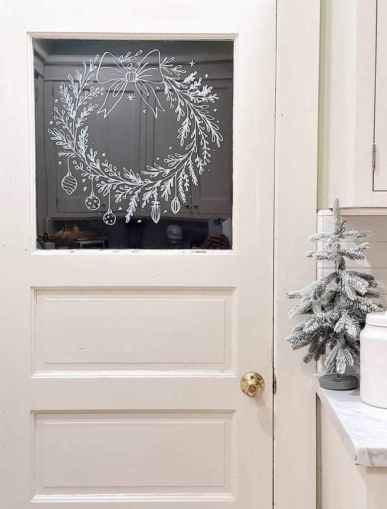 White door with a glass pane decorated with a hand-drawn wreath and ornaments; small artificial frosted tree on a nearby white countertop.