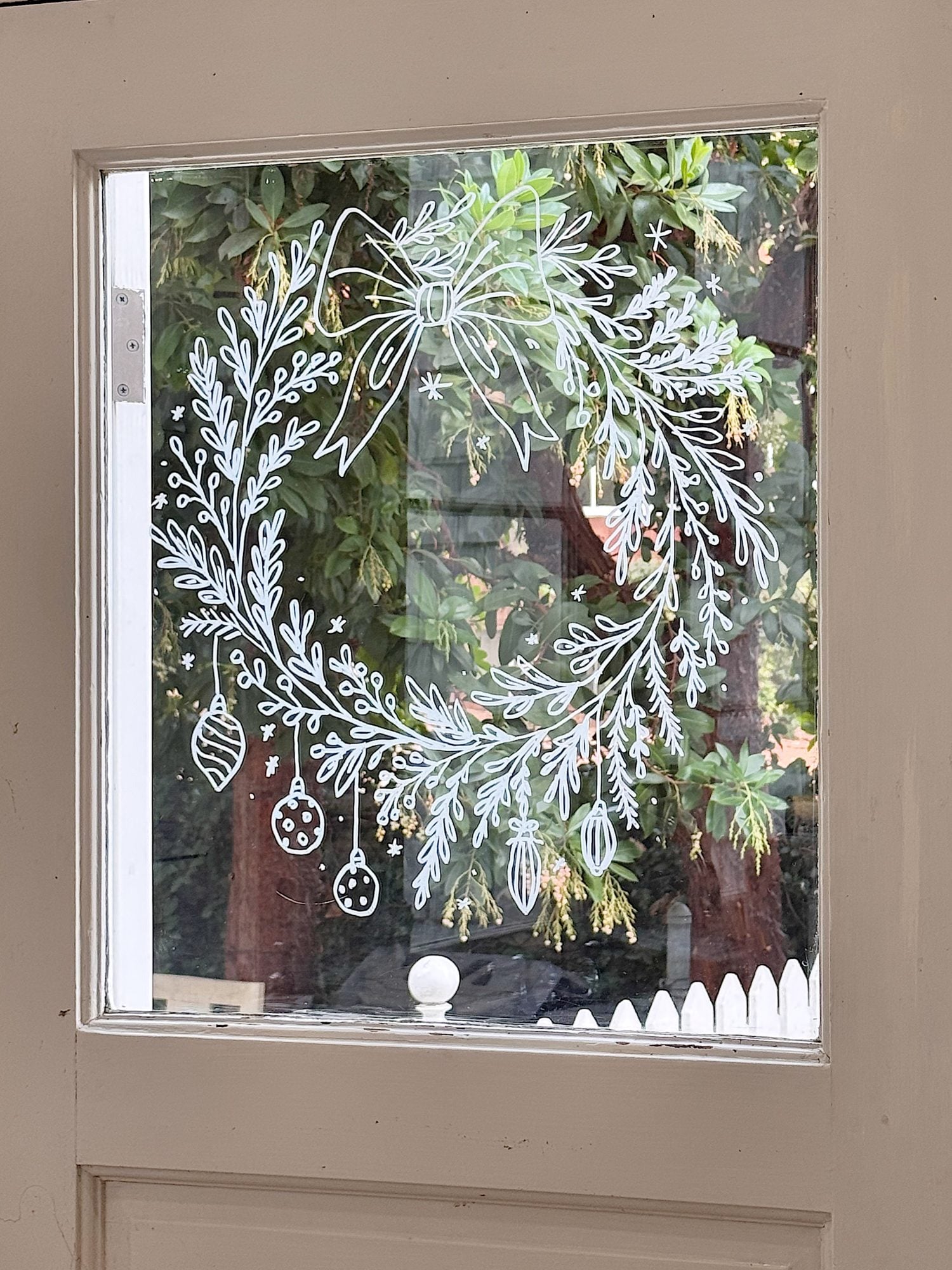 White marker drawing of a holiday wreath with ornaments and a bow on a glass door, with trees and a white picket fence visible outside.