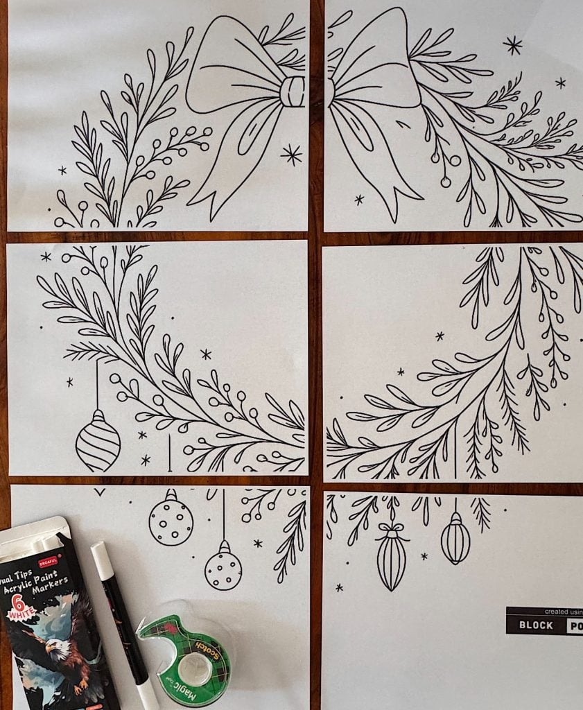 Six sheets of paper form a large outline drawing of holiday greenery and ornaments, with markers and tape positioned nearby on a wooden surface.