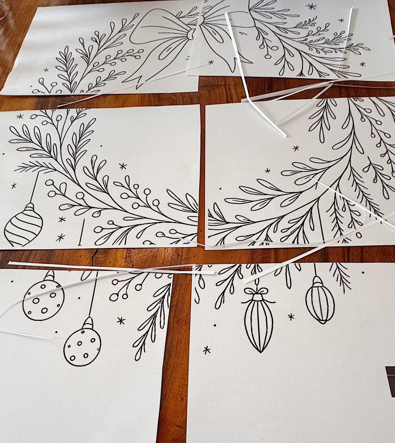 Four pieces of white paper with black line drawings of holiday ornaments and foliage are arranged on a wooden surface, with cut strips of paper scattered around.