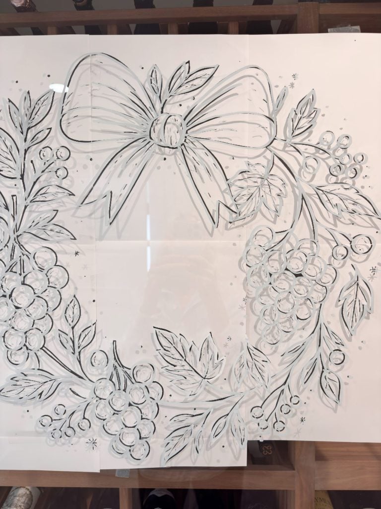 Black and white sketch of a wreath with leaves, berries, and a large bow at the top, drawn on a glass surface.