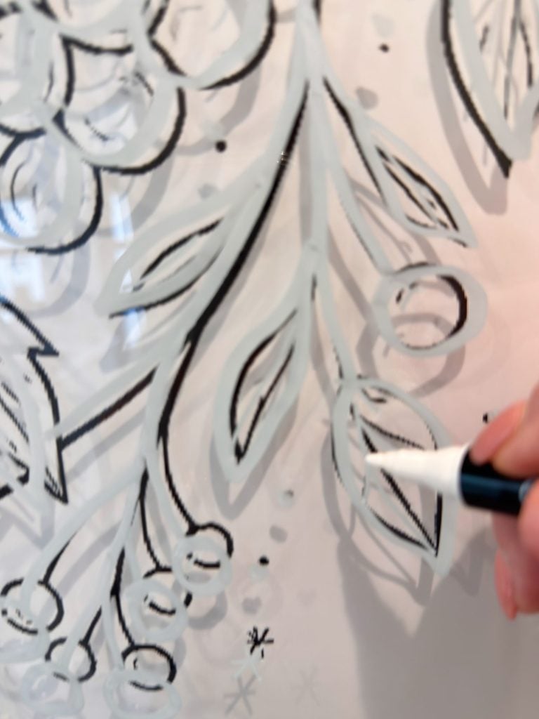 A hand holding a white marker draws leaves and berries on a glass surface with black and white lines.