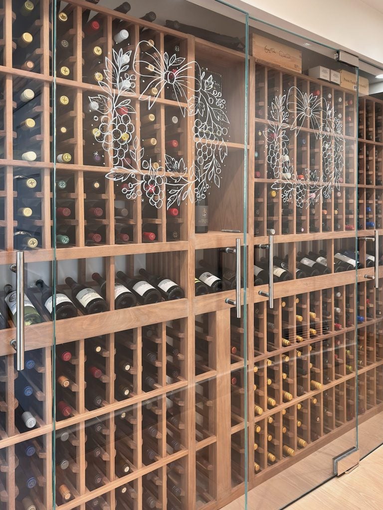 A wine cellar with wooden racks filled with wine bottles behind glass doors decorated with white grapevine illustrations.