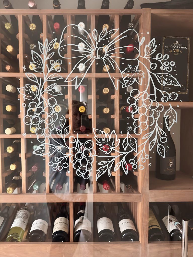 Wooden wine rack filled with assorted wine bottles, reflected glass door decorated with a white holiday wreath illustration.