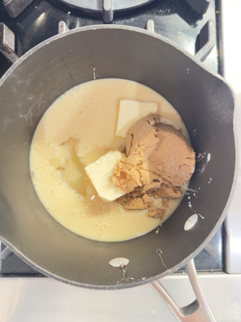 A saucepan on a stove contains brown sugar, butter, and condensed milk, partially mixed together.