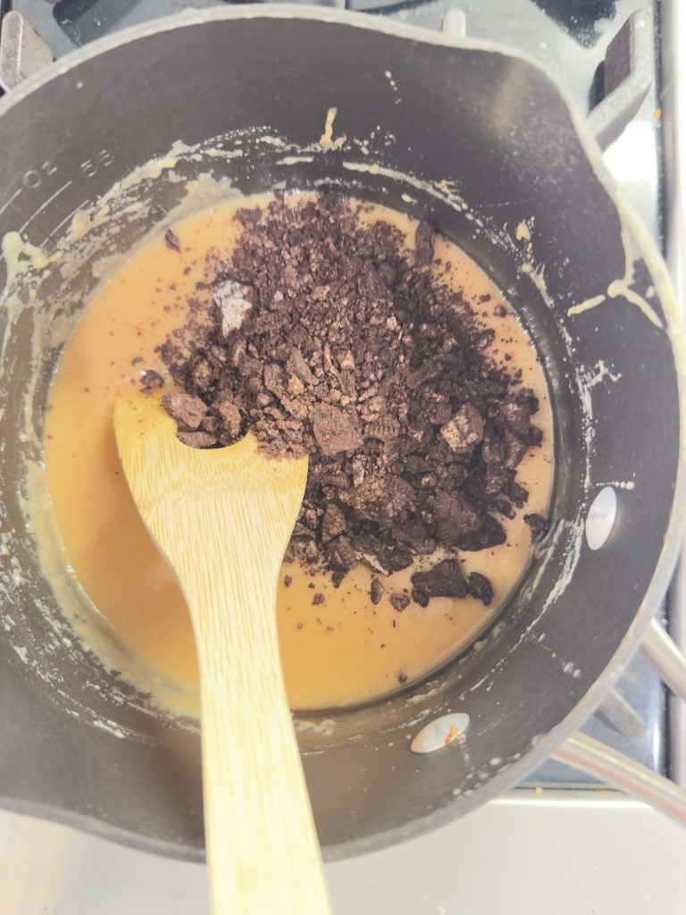 A saucepan on a stove with a mixture of caramel-colored liquid and crushed chocolate, being stirred with a wooden spoon.