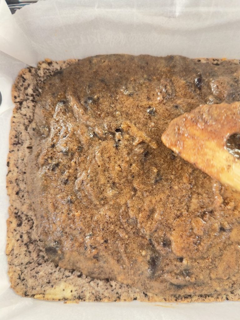 A close-up view of a freshly baked, rectangular cake with a golden-brown top, still in a parchment paper-lined baking pan. A wooden spatula is visible on the right side.
