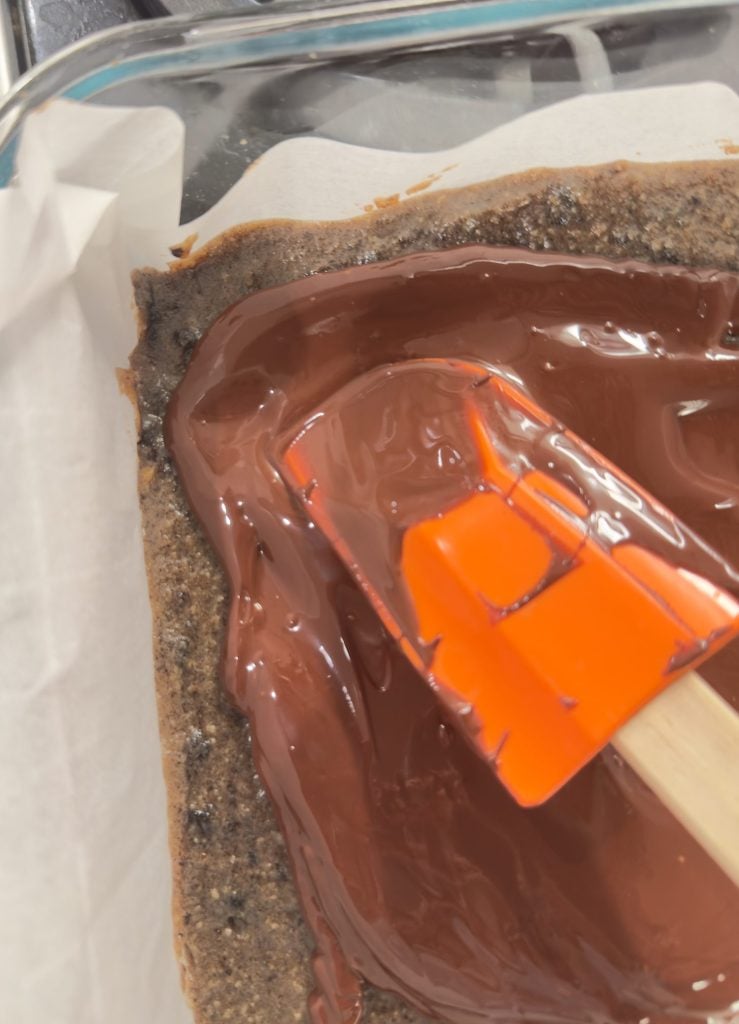 A close-up of a baking dish with a crumbly baked base, topped with melted chocolate being spread with an orange spatula.