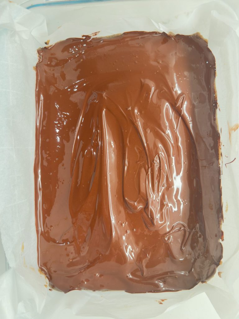 A rectangular pan lined with parchment paper holds a dessert topped with a smooth layer of glossy chocolate.