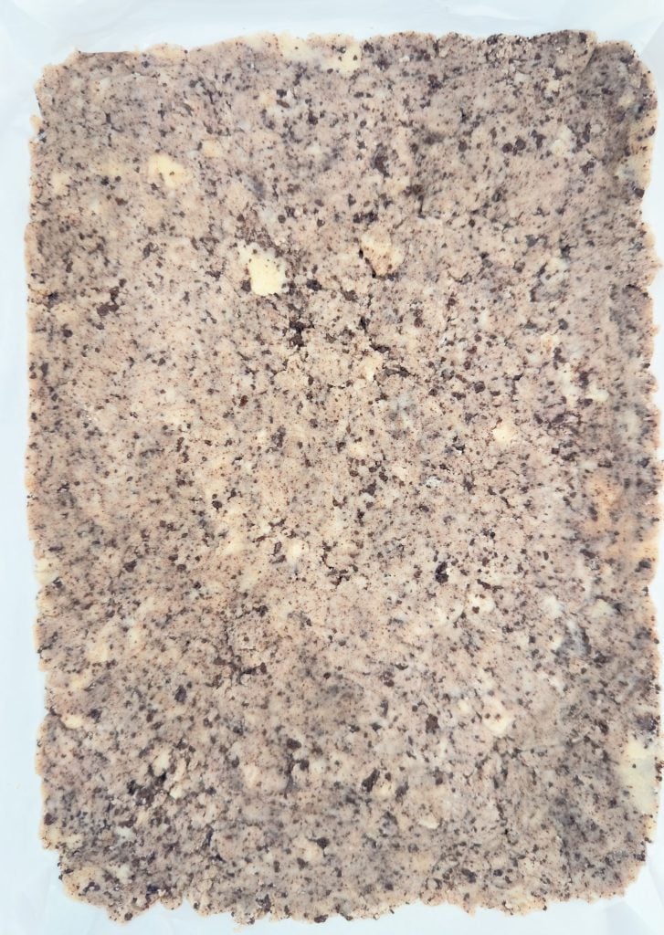 A rectangular sheet of light brown, crumbly dough with darker specks, pressed flat on parchment paper.