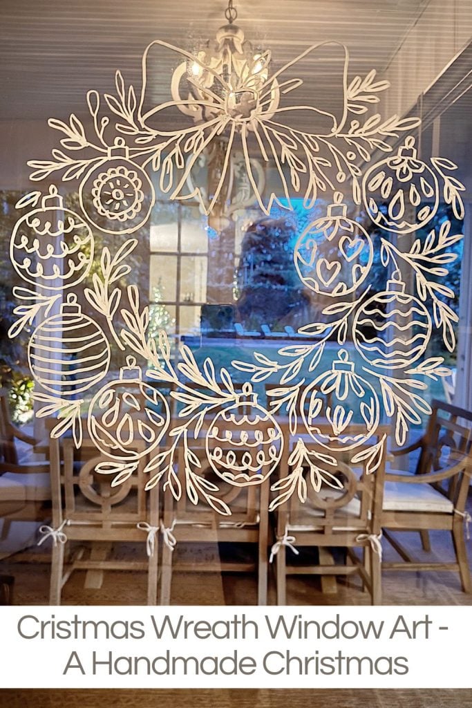 White paper cutout of a Christmas wreath with ornaments is displayed on a glass window; wooden chairs and chandelier are visible through the glass in the background.