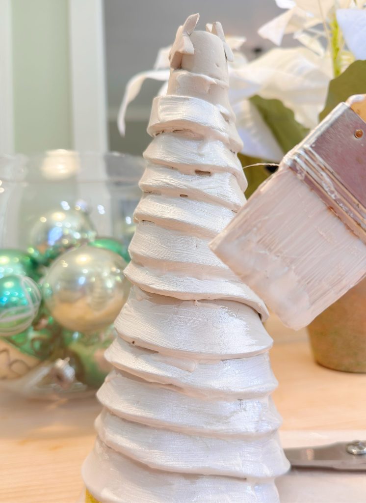 A hand uses a paintbrush to apply white paint to a spiral-wrapped cone, with green and silver ornaments in the blurred background.