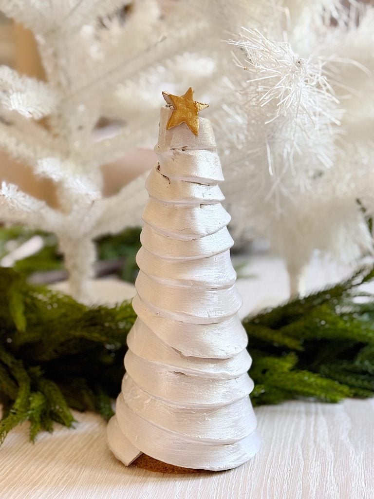 A white spiral Christmas tree decoration with a gold star topper sits on a table, surrounded by green and white artificial foliage.
