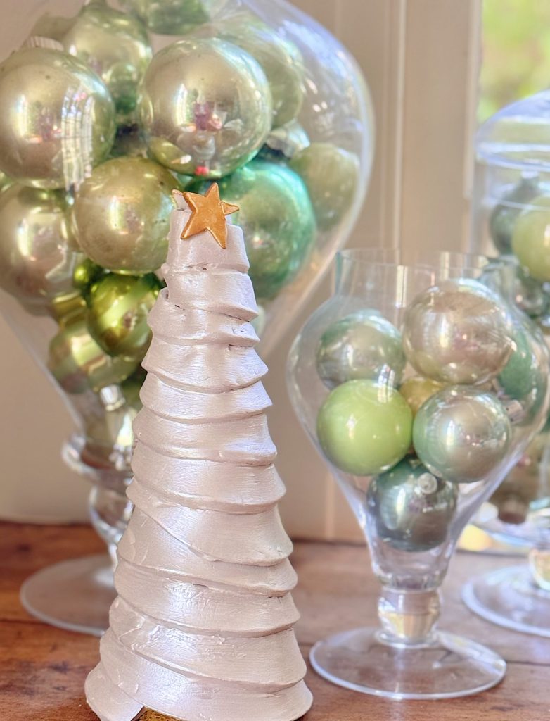 A white spiral Christmas tree decoration with a gold star stands in front of glass vases filled with green and silver ornaments.
