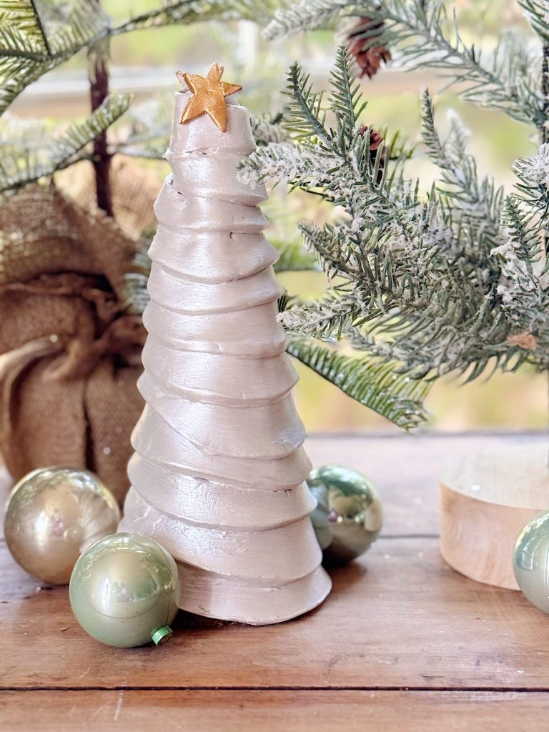 A ceramic Christmas tree with a gold star topper is displayed on a wooden surface, surrounded by green ornaments and pine branches.