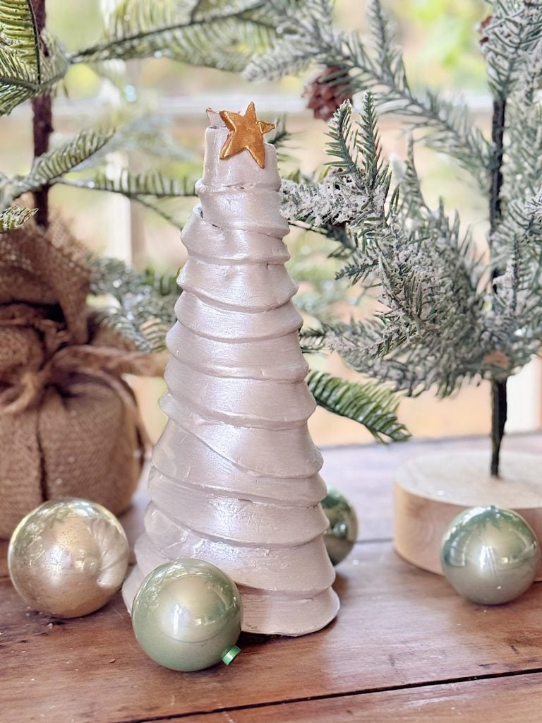 A white, spiral-shaped decorative Christmas tree with a gold star topper is displayed on a wooden table, surrounded by green ornaments and artificial pine branches.