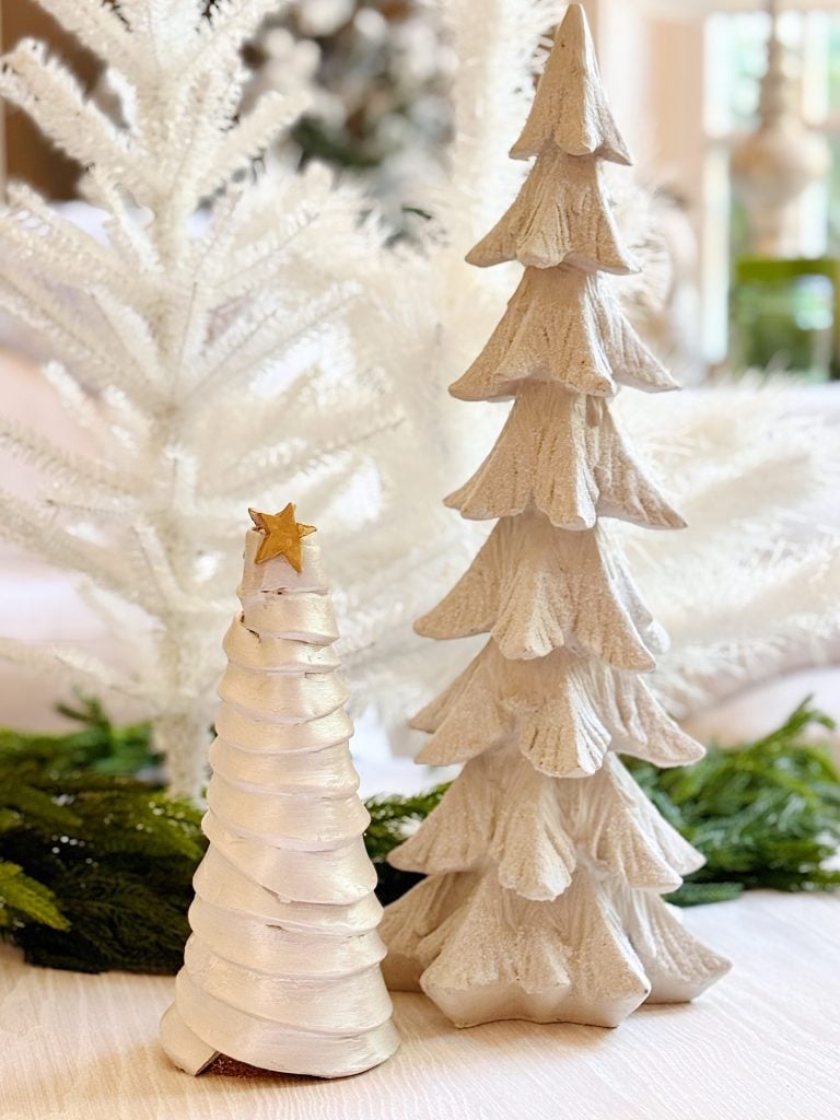 Two decorative white Christmas trees, one small with a gold star on top and one tall, are displayed with green foliage in the background.