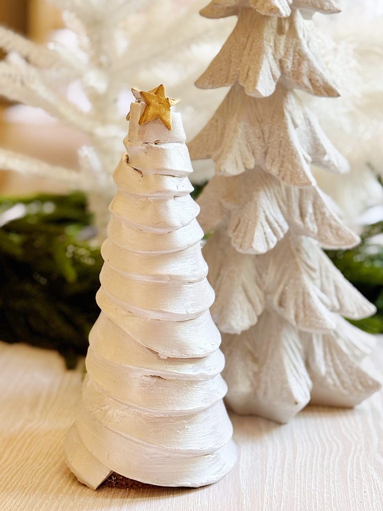 Two white, spiral-textured Christmas tree ornaments stand on a light wood surface, one topped with a small gold star. Greenery is visible in the background.