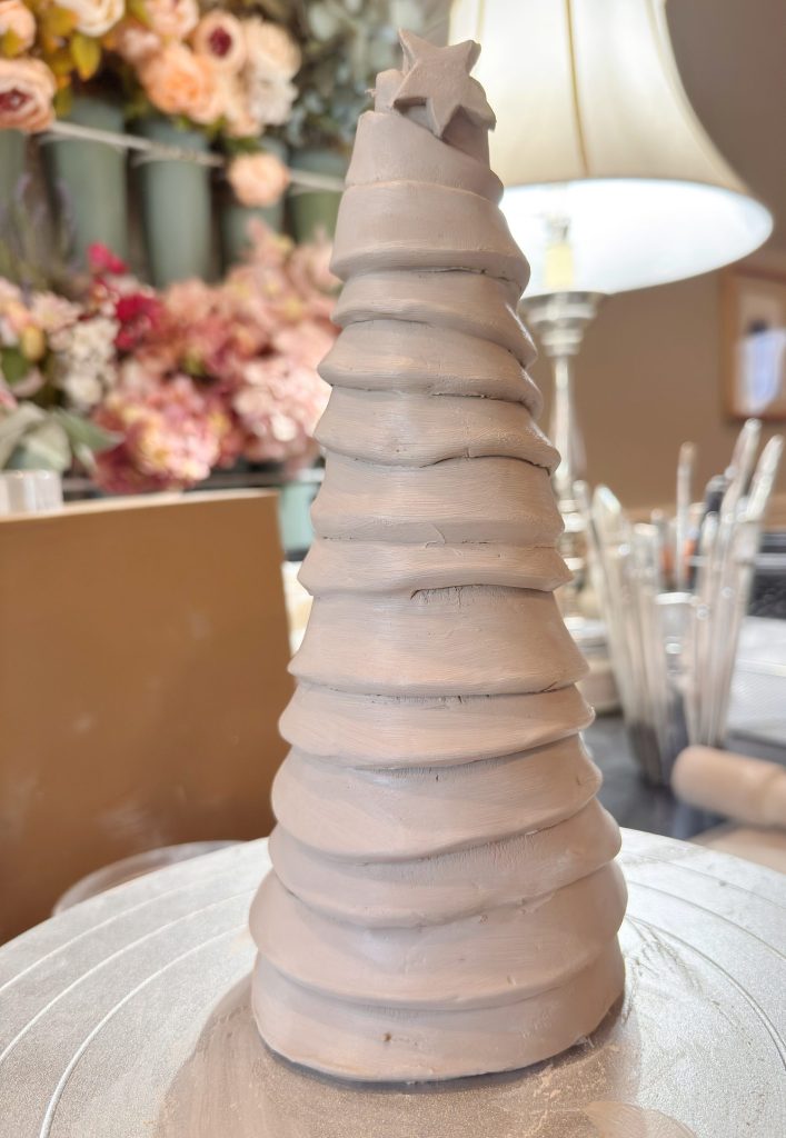 A cone-shaped clay sculpture resembling a stylized Christmas tree with layered ridges and a star on top, placed on a work surface with art supplies and flowers in the background.