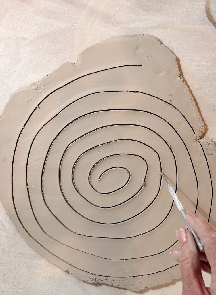 A hand uses a metal tool to cut a spiral pattern into a flat slab of clay.