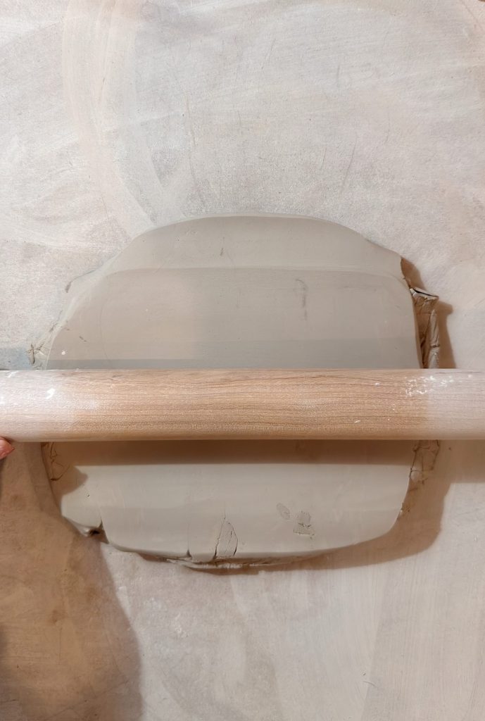 A hand uses a wooden rolling pin to flatten a slab of grey clay on a light-colored surface.