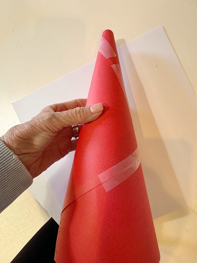 A hand holds a red paper cone secured with clear tape, with a sheet of white paper on a light-colored surface in the background.