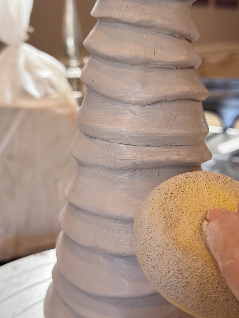 A hand uses a sponge to smooth the surface of a ridged, gray clay sculpture.
