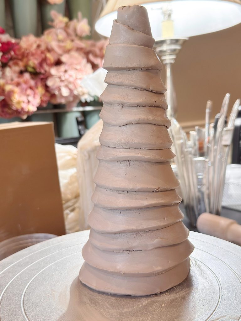 A smooth, spiral-shaped clay sculpture stands upright on a circular surface, with flowers and a lamp in the background.