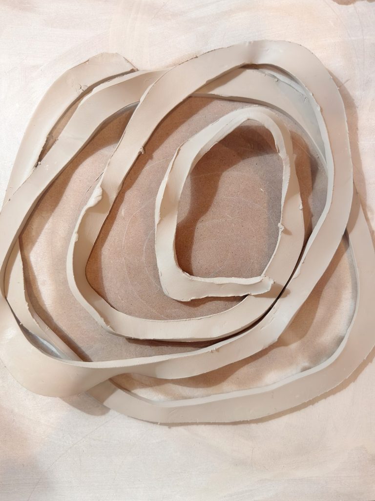 Several irregular, flat strips of light-colored clay are arranged in overlapping, looped shapes on a smooth, neutral surface.