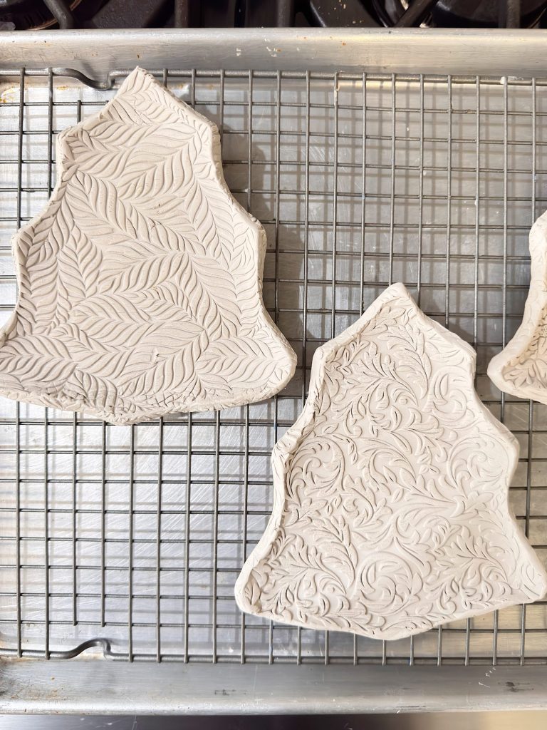 Two pieces of raw clay shaped like irregular polygons with embossed leaf and floral patterns rest on a wire rack over a baking sheet.