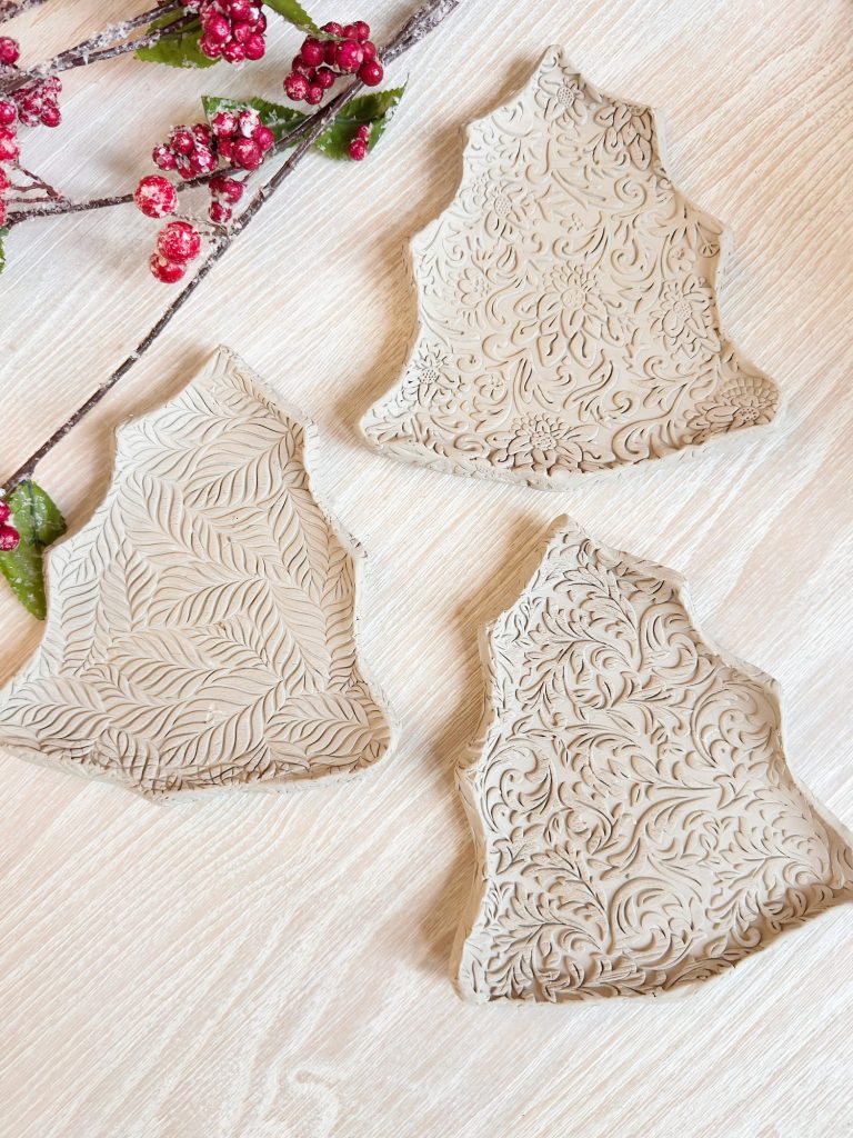 Three clay trays shaped like Christmas trees with intricate leaf and floral patterns are displayed on a light wooden surface next to a branch with red berries.