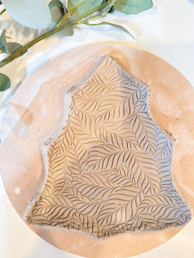 A piece of clay shaped like a Christmas tree with a leaf pattern texture, placed on a round beige surface with green foliage nearby.