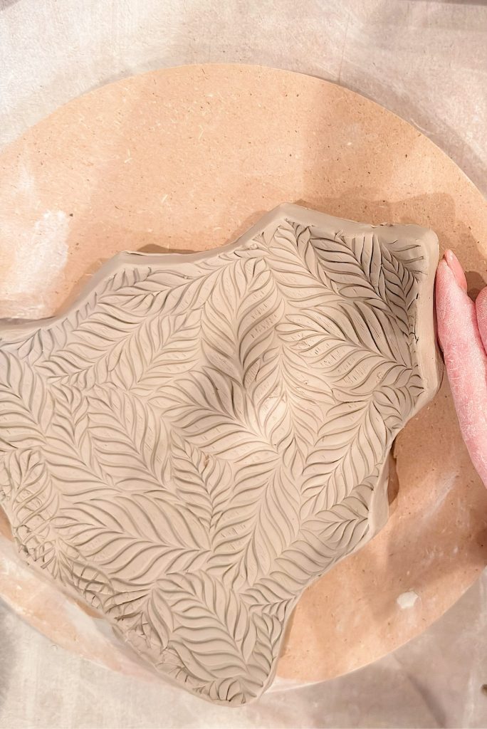 Clay slab with an embossed leaf pattern rests on a round wooden board; a hand in a pink glove is visible on the right side of the image.