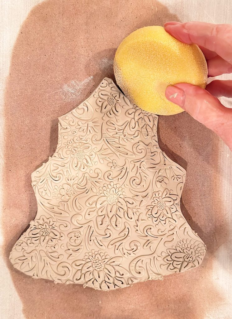 A hand holds a yellow sponge above a piece of clay with an ornate floral pattern embossed on its surface, resting on a sheet of brown paper.