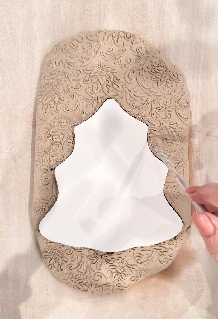 A hand uses a pottery tool to cut around a paper template on a slab of textured clay with a floral pattern.