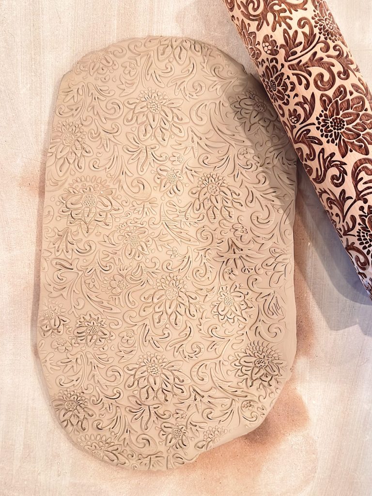 A slab of clay with an intricate floral pattern embossed by a decorative rolling pin placed beside it.