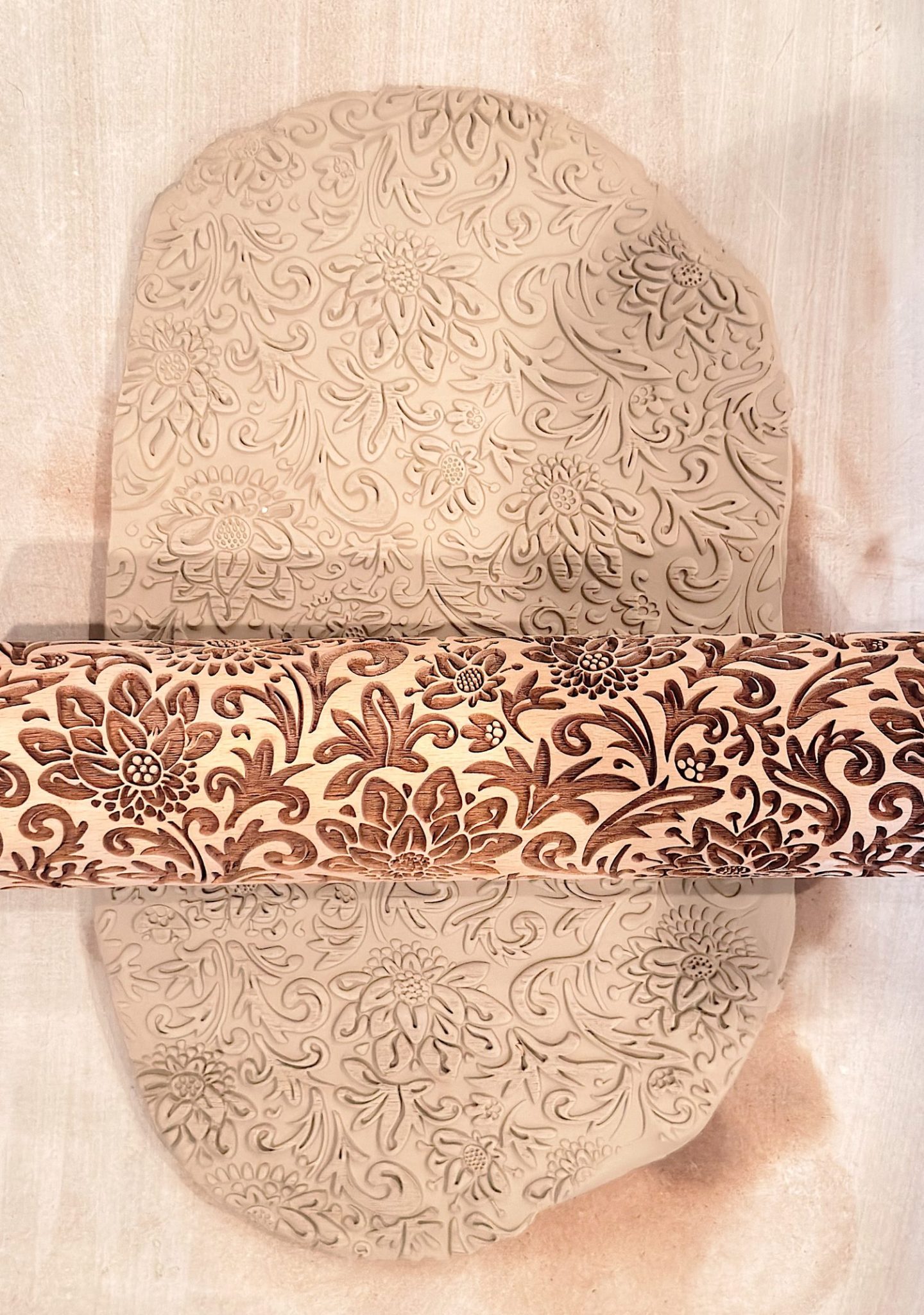 A rolling pin with a floral pattern embosses a sheet of clay, transferring the intricate design onto the surface.
