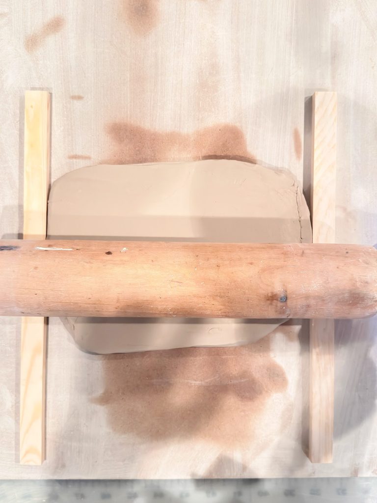 A slab of clay is being flattened with a rolling pin, guided by two wooden slats on either side.